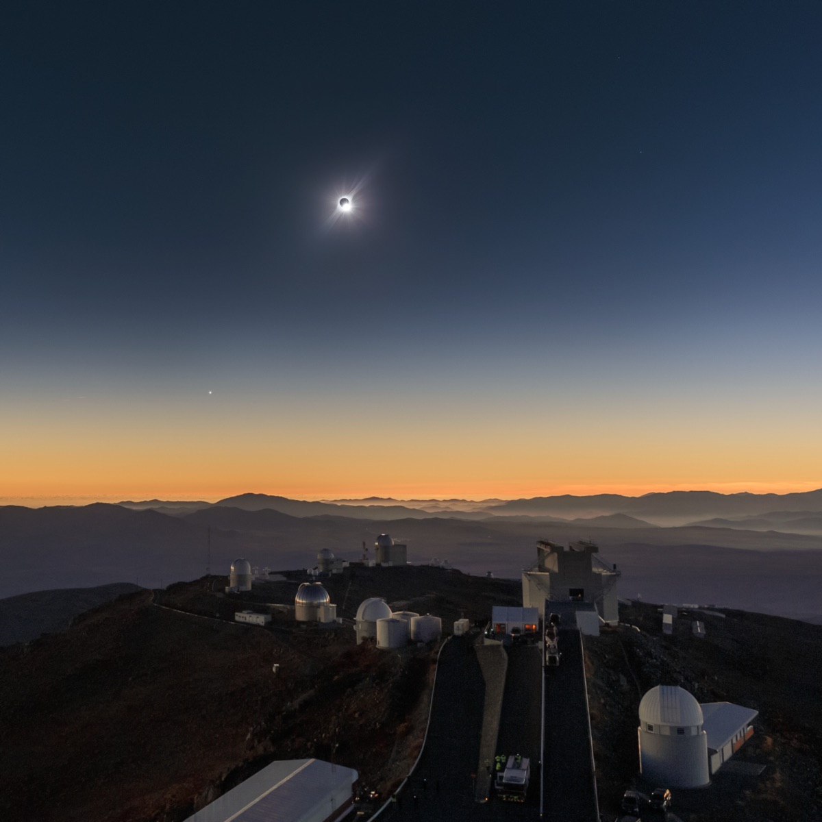 The 2019 total eclipse seen from the mountaintop La Silla Observatory: the corona in a darkened sky above telescope domes, twilight colours on every horizon