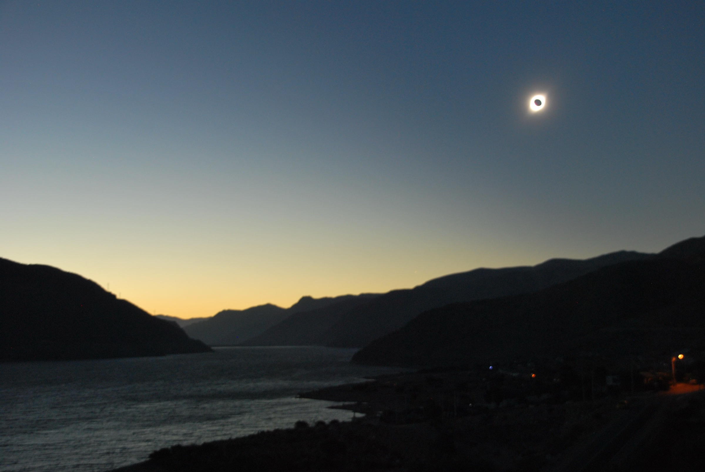 The totally eclipsed Sun, corona and diamond ring, hanging low over a mountain-ringed reservoir at deep twilight in Chile, 2019