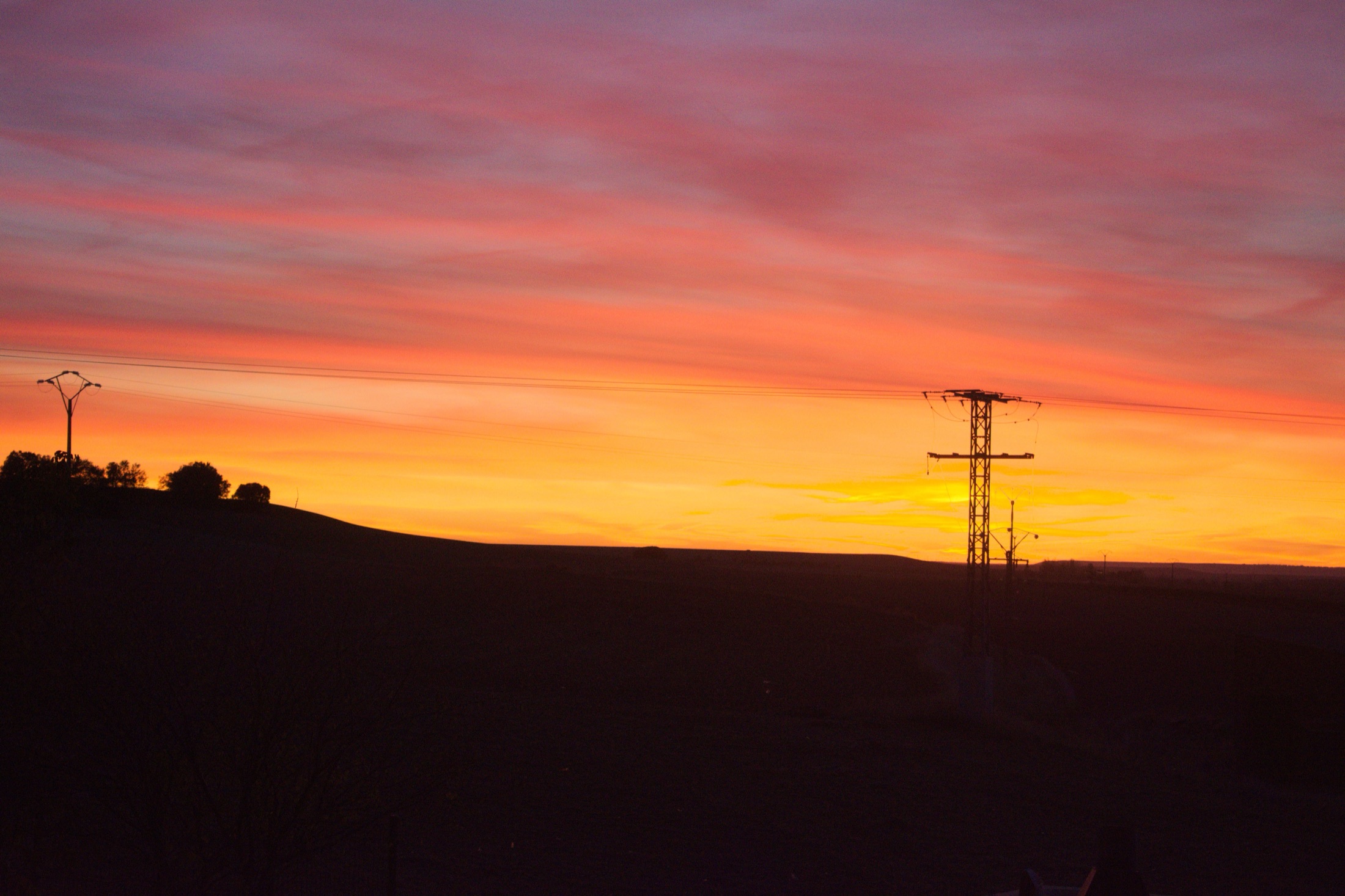 A dead-flat Castilian plain silhouetted against an enormous red-and-orange sunset sky, a single power pylon and a low rise the only features