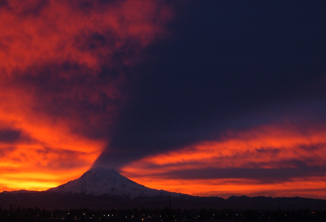 Mount Rainier at sunrise casting a dark shadow cone upward onto the cloud layer above it, against an orange sky