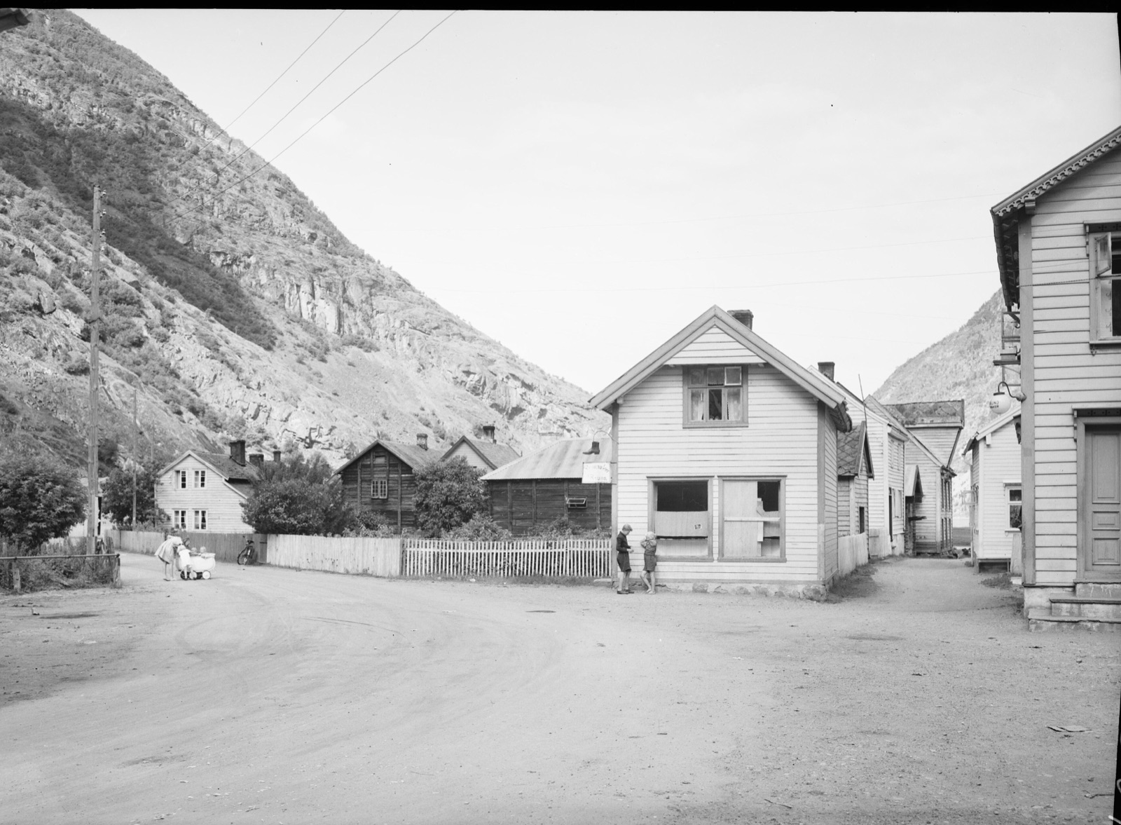 A dirt street in Lærdalsøyri lined with white clapboard houses, children with a pram at the roadside, and the bare rock wall of the fjord side rising directly behind the roofs
