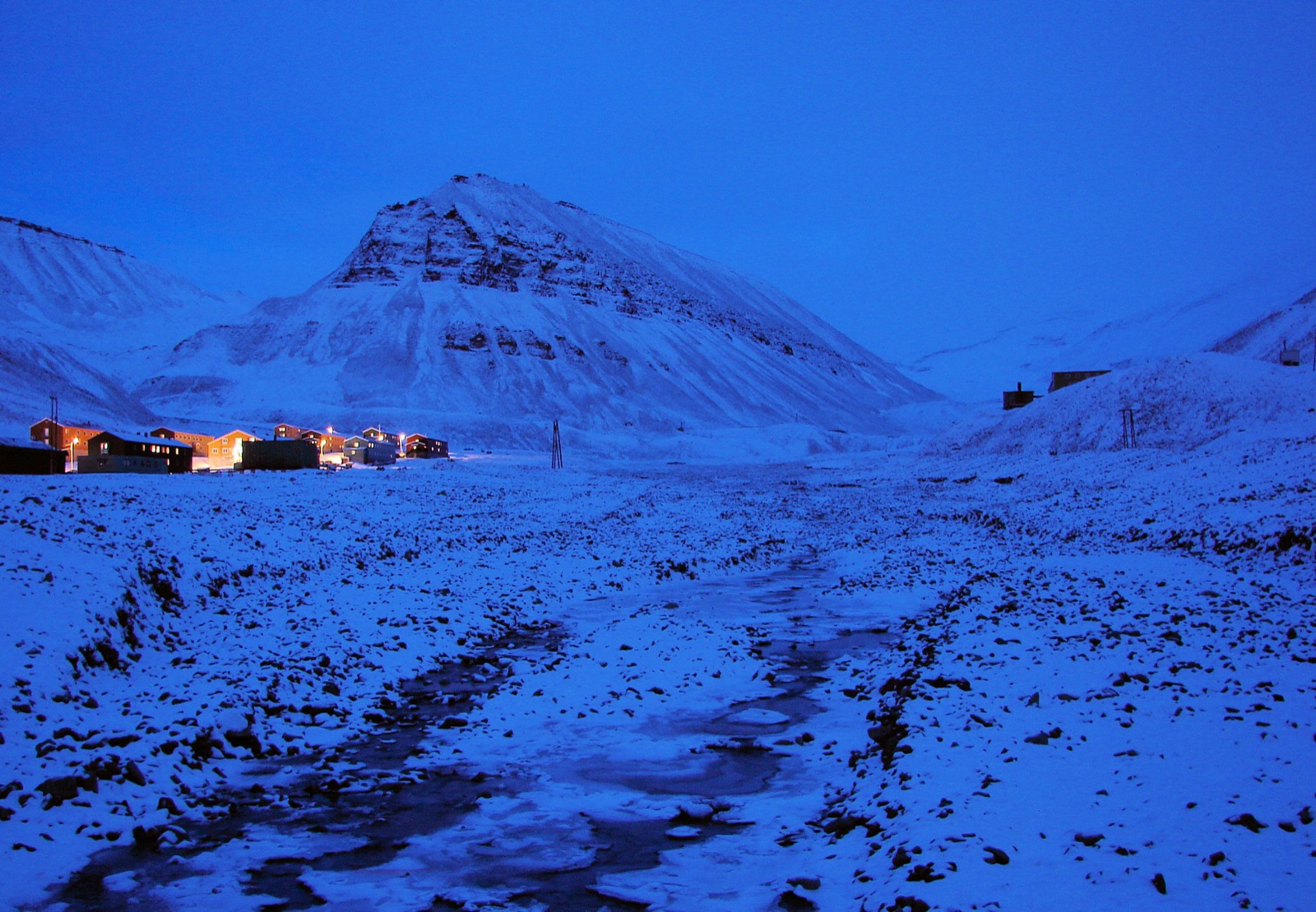 Longyearbyen in polar twilight: a row of houses with warm lit windows on a snowfield beneath a dark pyramidal mountain, the whole scene drenched in deep blue
