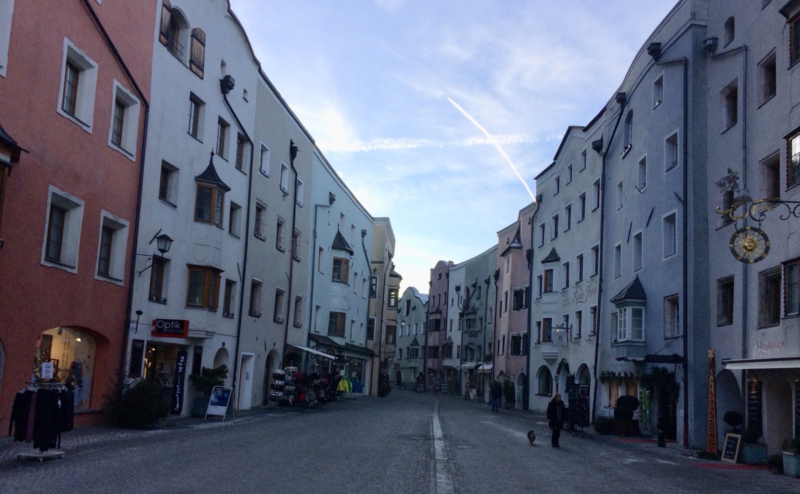 Rattenberg's main street two days after the winter solstice: tall painted houses on both sides, shop lights on, a woman walking a dog, the entire street floor in flat shade under a bright blue sky