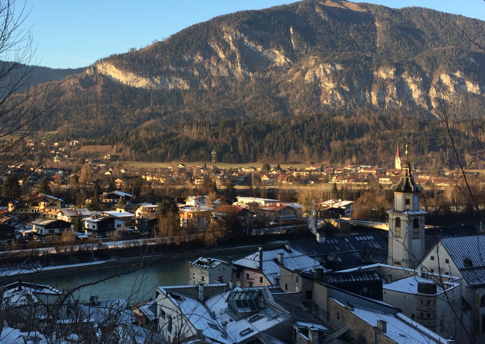 Rattenberg in Tyrol on a December afternoon: the town's snow-covered roofs in the foreground lie in cold blue shade while the houses on the far bank of the Inn and the mountain above them are still lit warm gold by the low sun